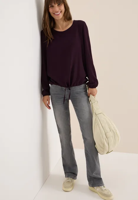 Frau mit lila Bluse, grauer Jeans und weißer Tasche lehnt lächelnd an der Wand.
